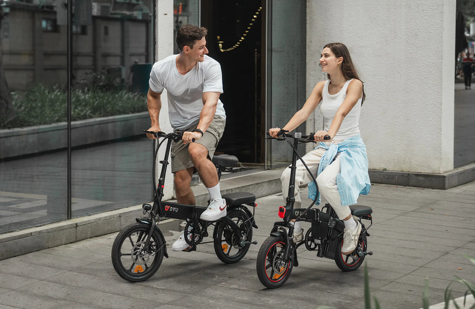 A man and a woman riding electric bikes on a city sidewalk, both smiling and looking at each other. The man wears a white t-shirt and shorts; the woman wears a white top and pants with a blue jacket tied around her waist.