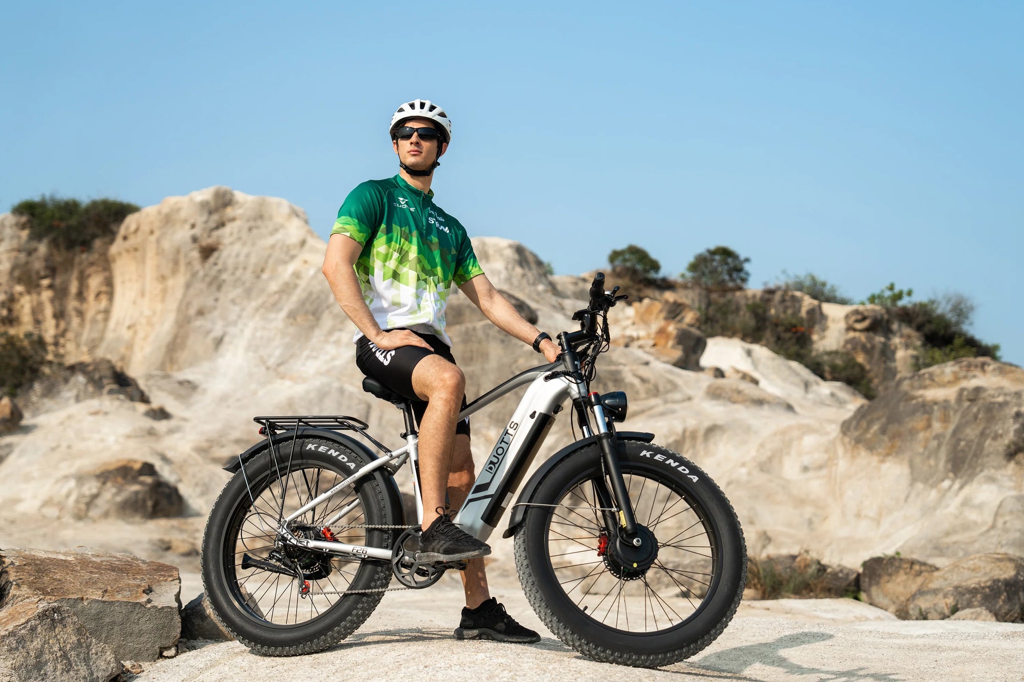 Man wearing helmet and sunglasses standing with a black and white fat tire electric bike on rocky terrain under clear blue sky