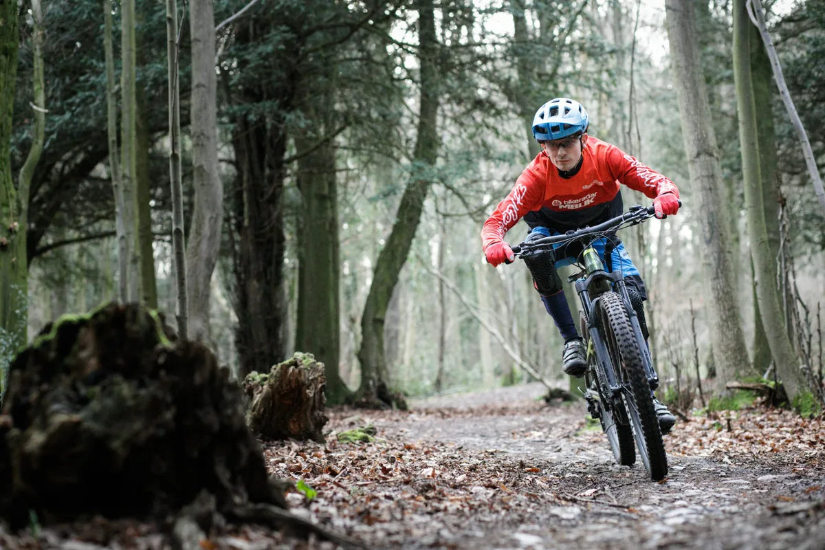 Man wearing blue helmet and red jacket riding a mountain bike on a forest trail covered with leaves and dirt.