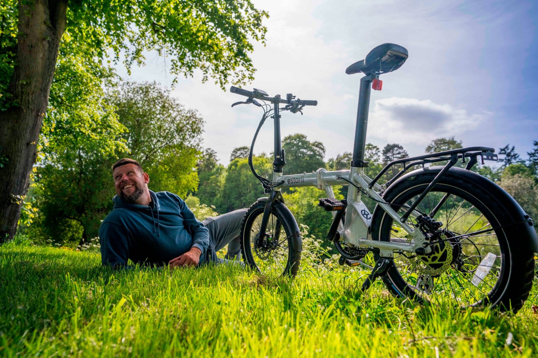 A man relaxes on the grass under a tree, smiling, with a folding bicycle parked beside him on a sunny day in a green, park-like setting.