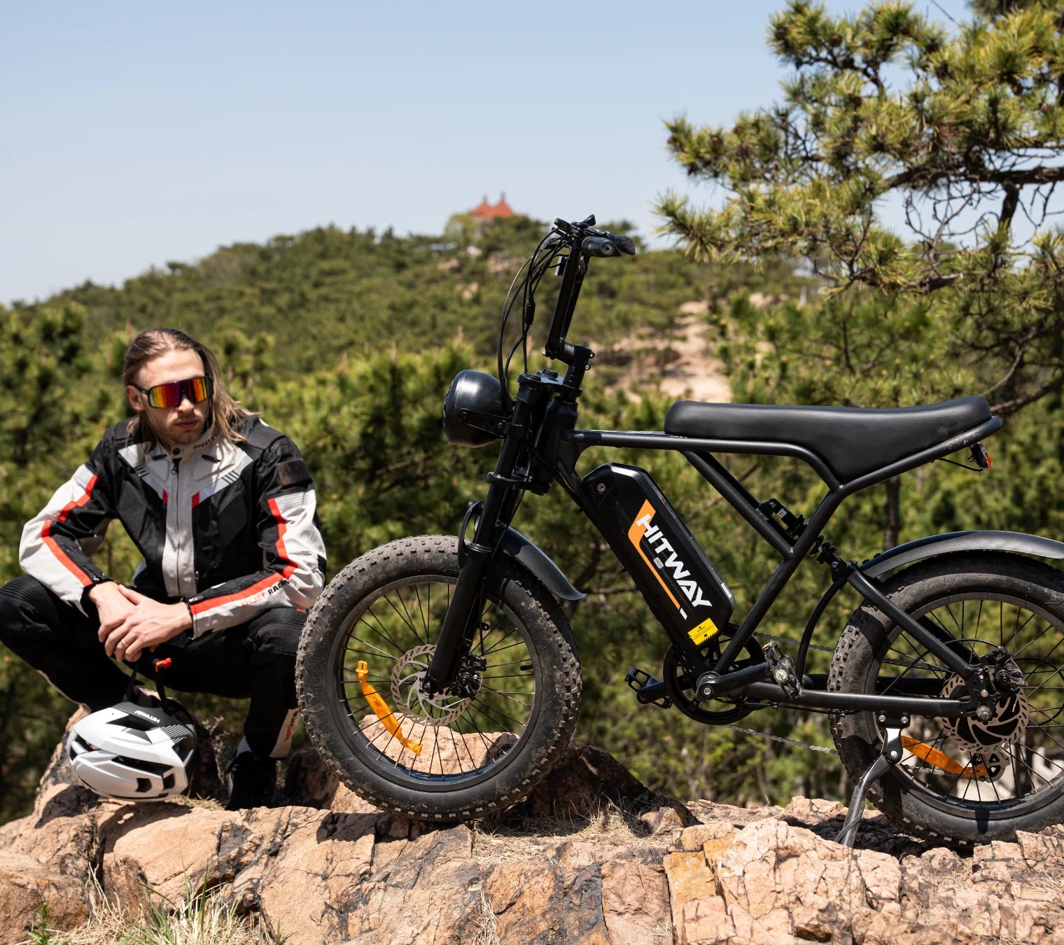 Man in motorcycle jacket sitting on a rock next to a black electric bike on a trail with trees and hills.