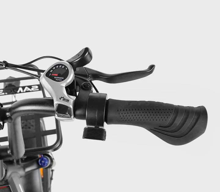 Close-up of a black bicycle handlebar featuring a textured grip, brake lever, gear shifter with visible numbers, and part of a metal basket in the background against a light gray backdrop.