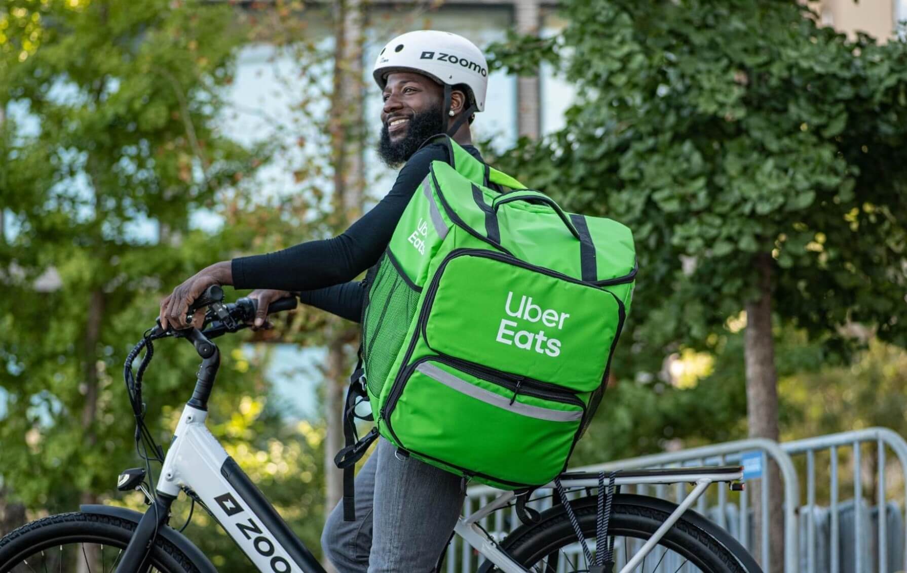 Man wearing a white bike helmet and black shirt riding a white bike with a large bright green Uber Eats delivery bag on his back outdoors