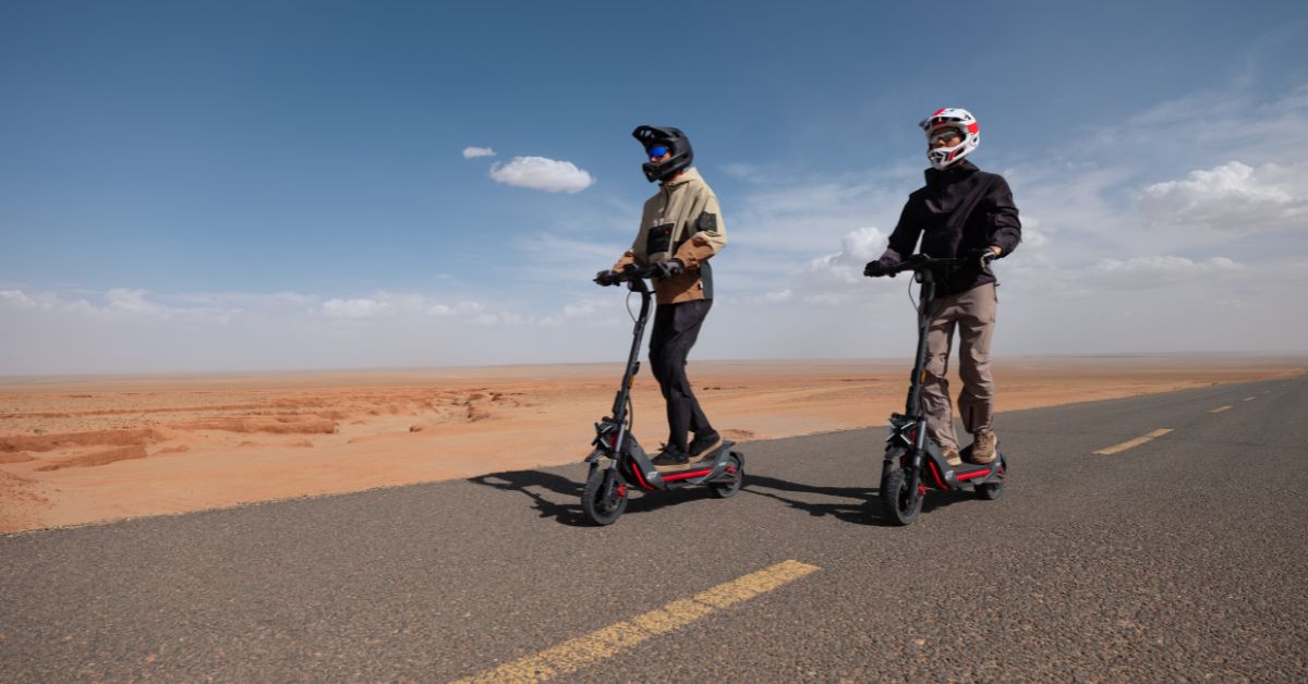 Two people wearing helmets ride electric scooters on an empty road through a vast, arid desert landscape under a bright, partly cloudy sky.