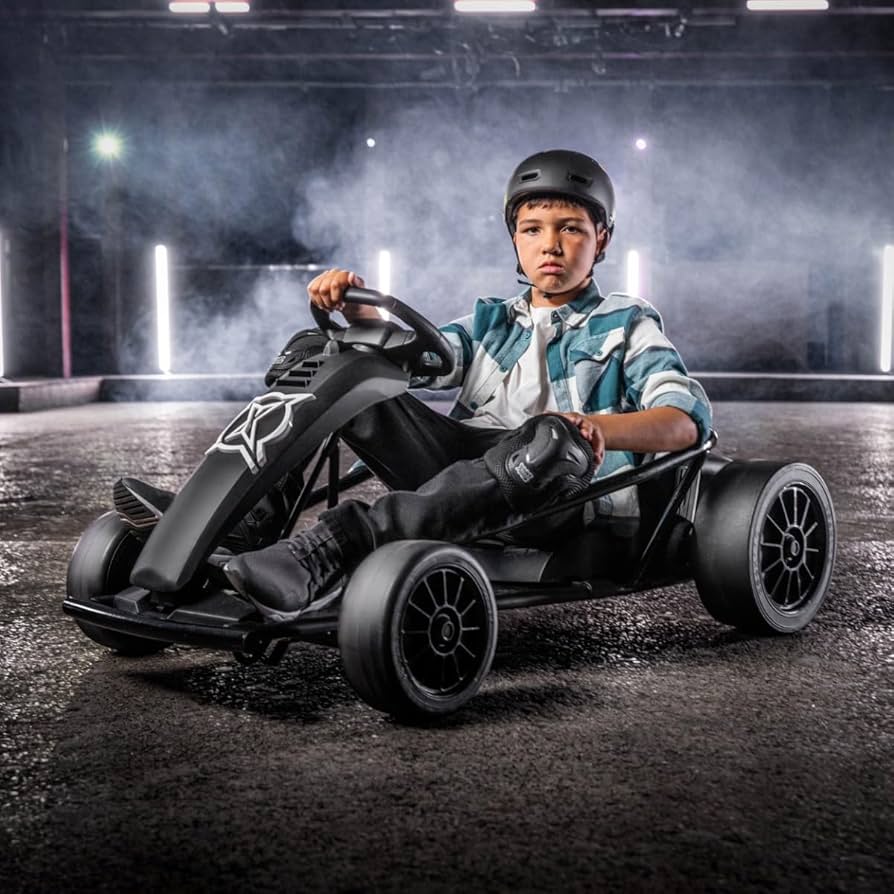 Boy wearing helmet and knee pads sitting on a black go-kart on a smoky racetrack with bright lights behind him