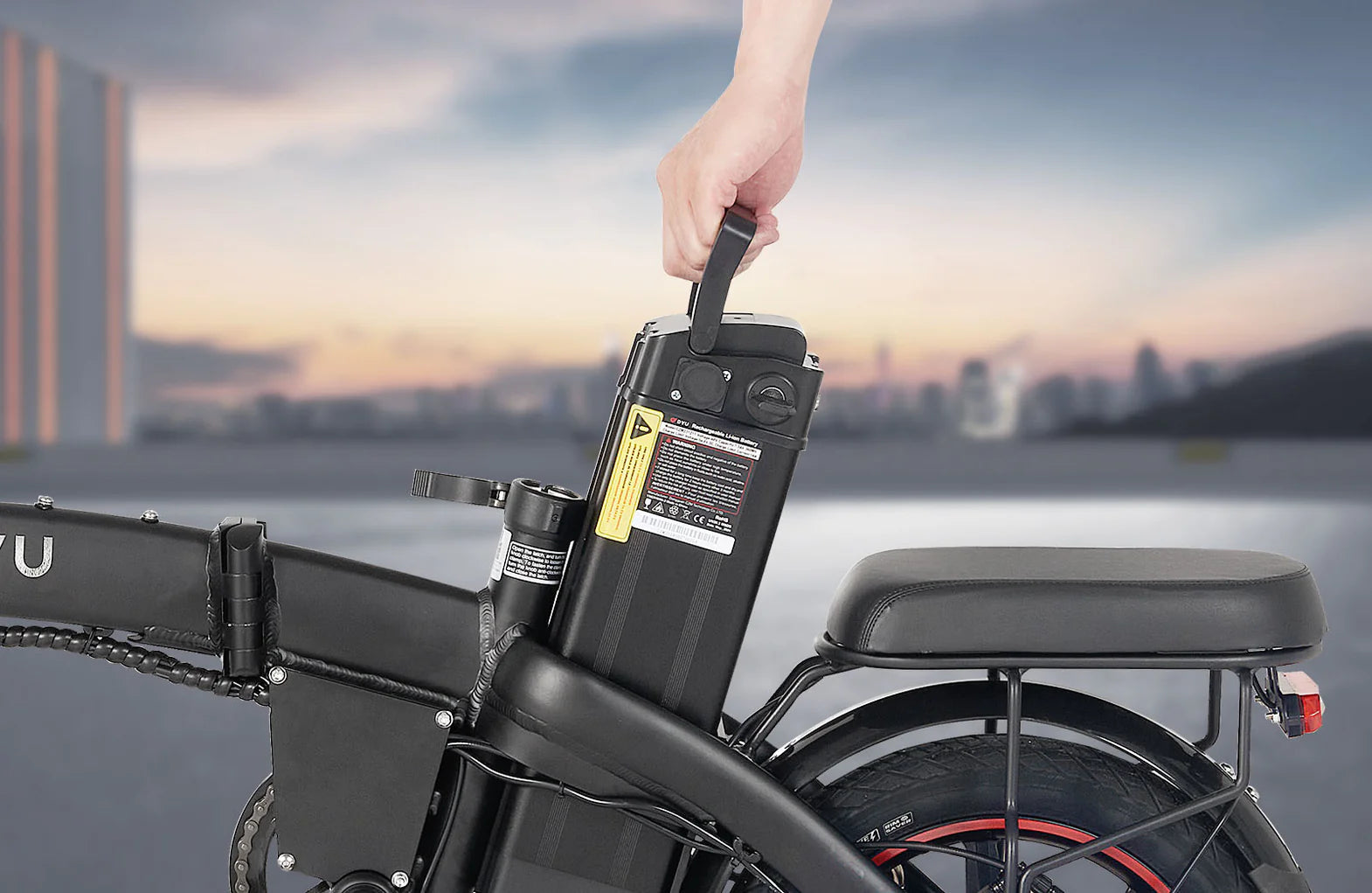 A hand lifts the removable battery from an electric bicycle, which is parked outdoors with a city skyline blurred in the background.