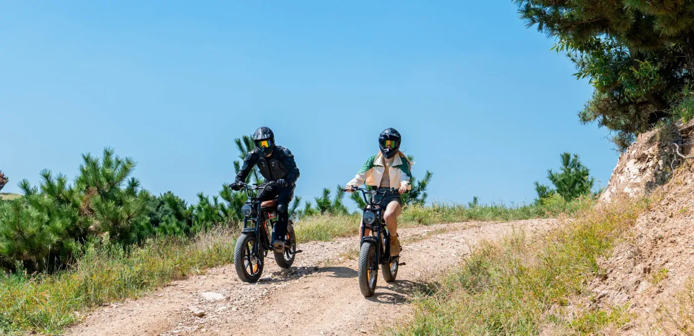 Two people wearing helmets riding electric bikes on a dirt trail surrounded by greenery under a clear blue sky.
