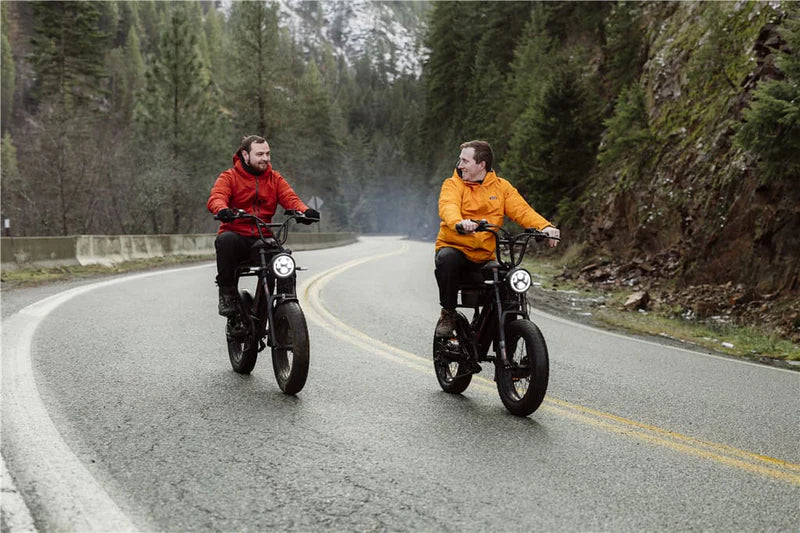 Two men wearing orange jackets riding electric bikes on a winding mountain road surrounded by trees.