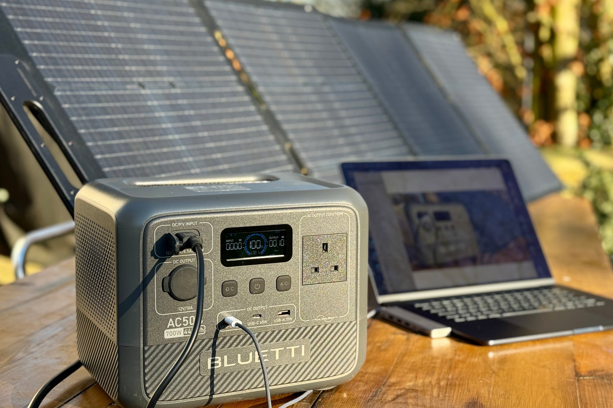 Portable Bluetti AC50 power station with cables connected sitting on a wooden table next to a laptop and solar panels outdoors