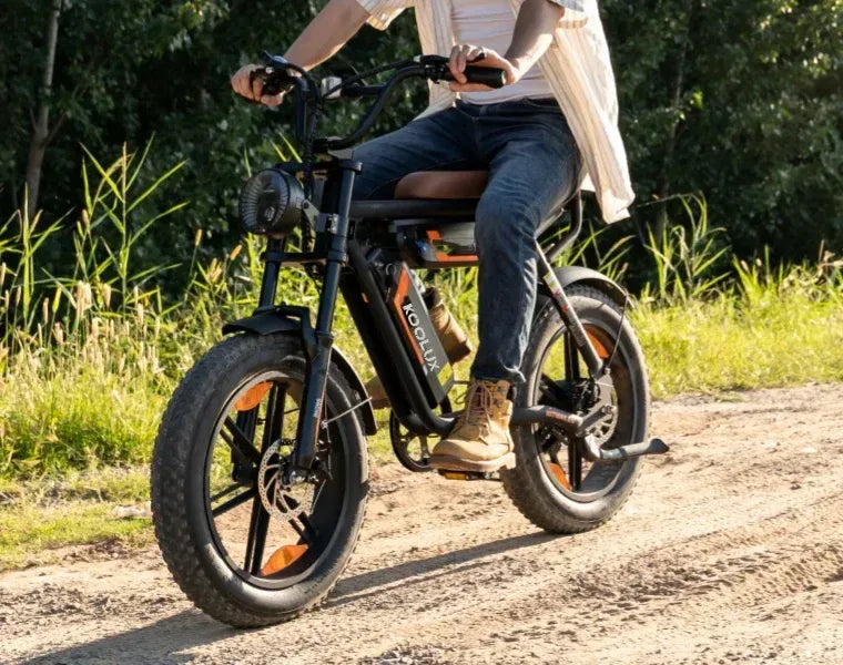 A person rides a black electric bike with thick tires on a dirt path, surrounded by green grass and trees. The rider wears jeans, boots, and a light shirt.