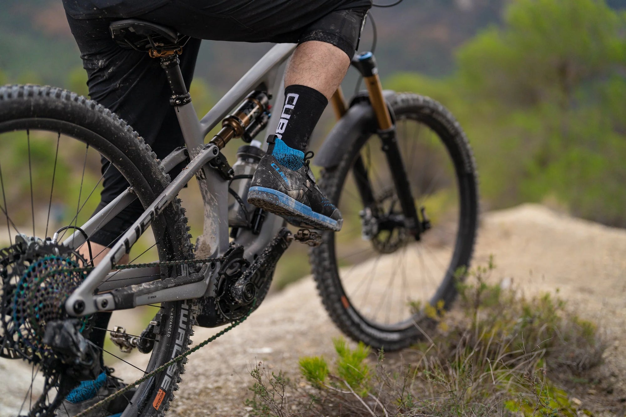 Close-up of a person pedaling a gray mountain bike on a dirt trail with greenery in the background