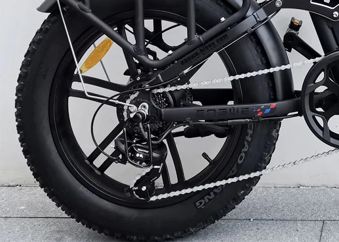 Close-up of a black bicycle’s rear wheel and drivetrain, showing the chain, gears, derailleur, disc brake, thick tire, and part of the black frame against a light gray background.