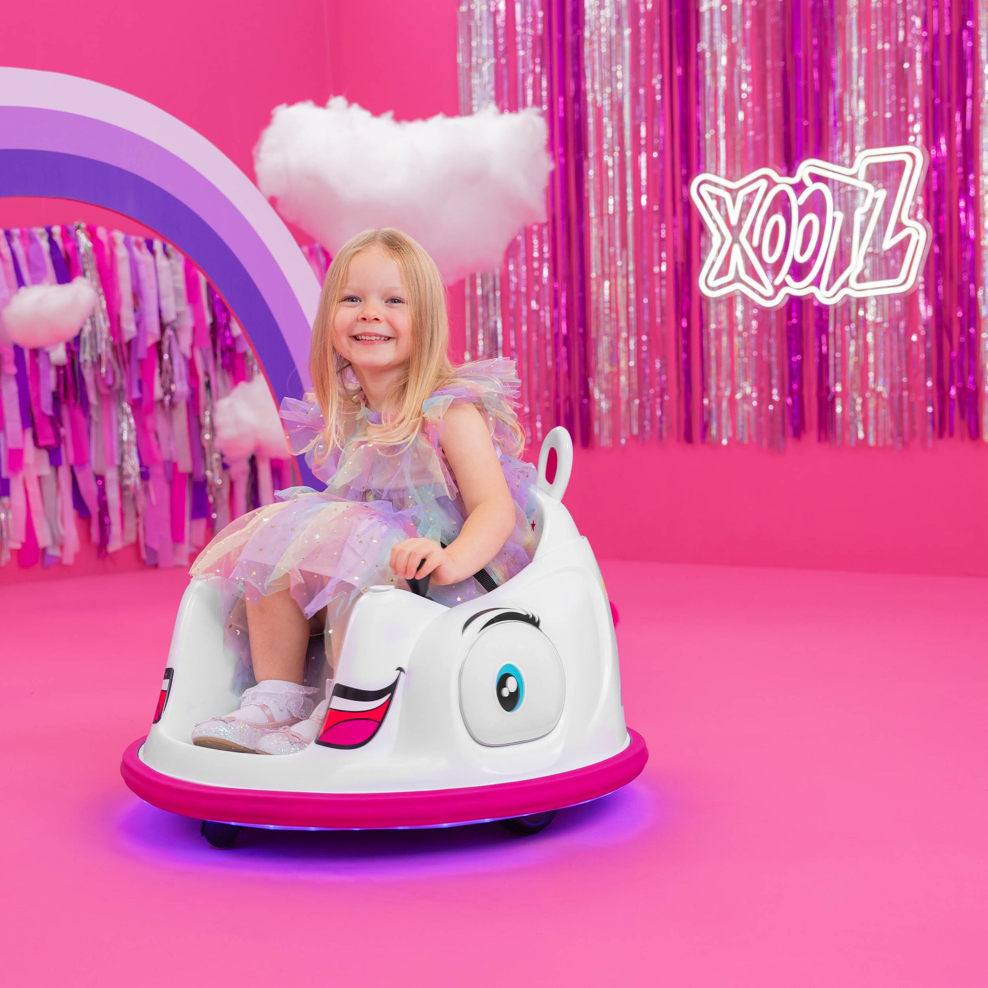 Young girl in a colorful dress riding a white cartoon-themed bumper car on a bright pink floor with decorations.
