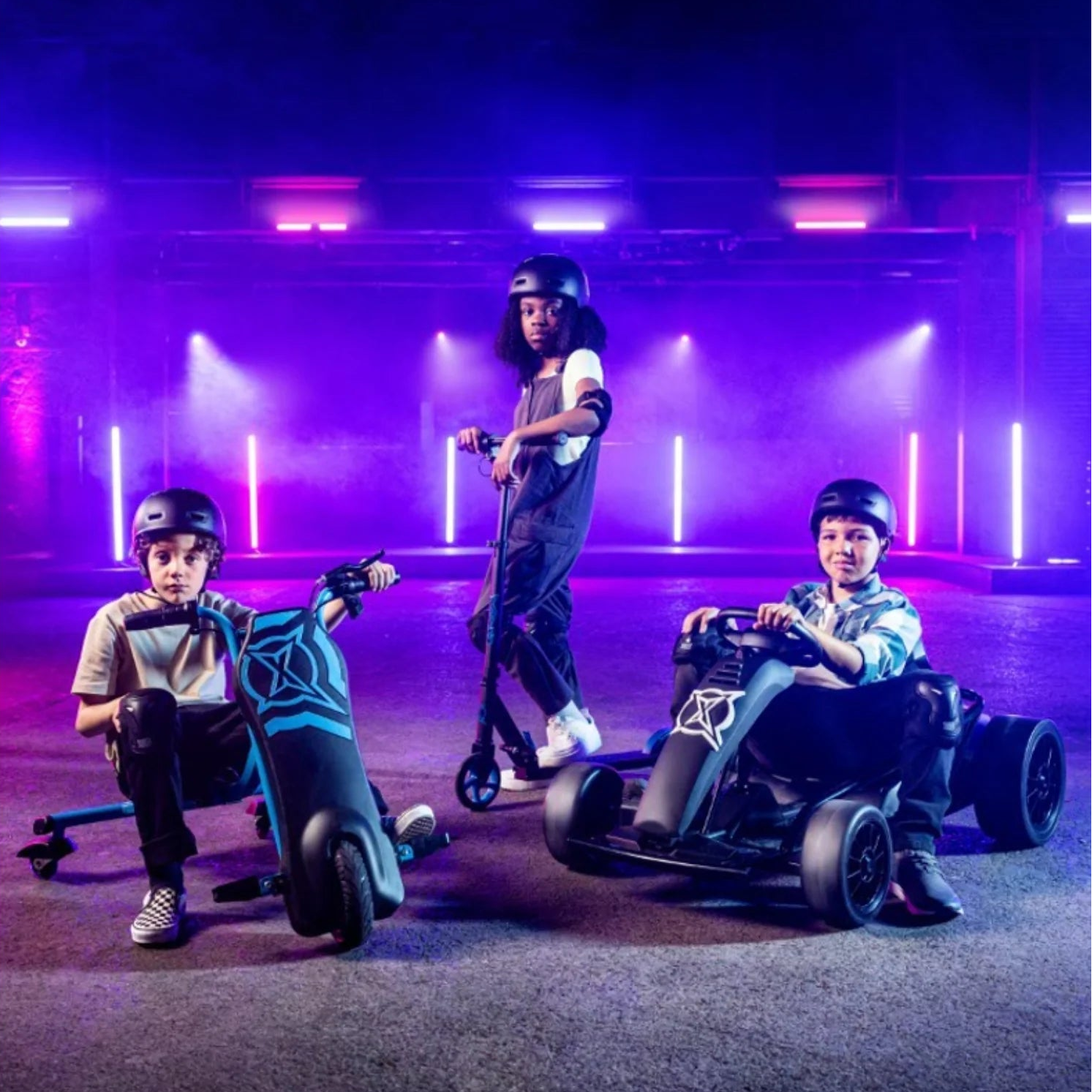 Three children wearing helmets posing with electric scooters and a go-kart in a neon-lit indoor space.