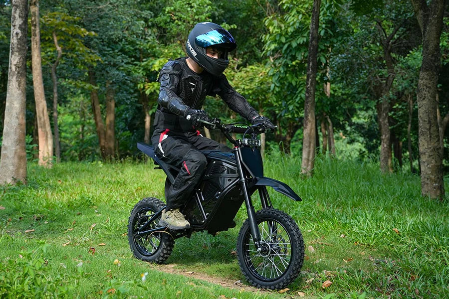 Person wearing black protective gear and helmet riding a black electric dirt bike on a grassy forest trail