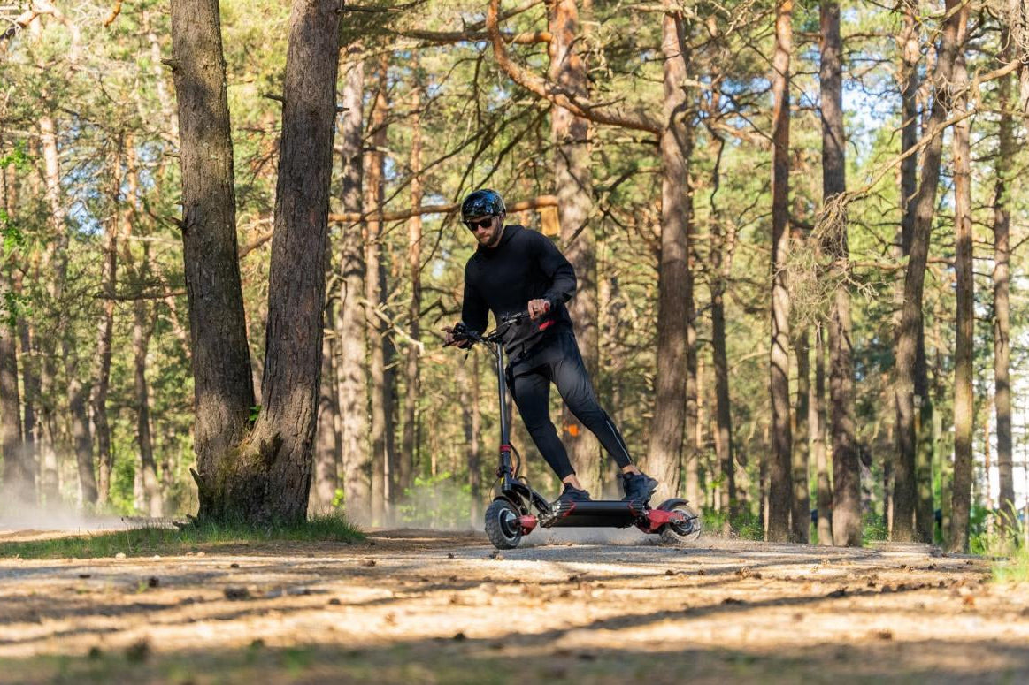 Man riding a black electric scooter on a forest trail surrounded by tall pine trees in daylight