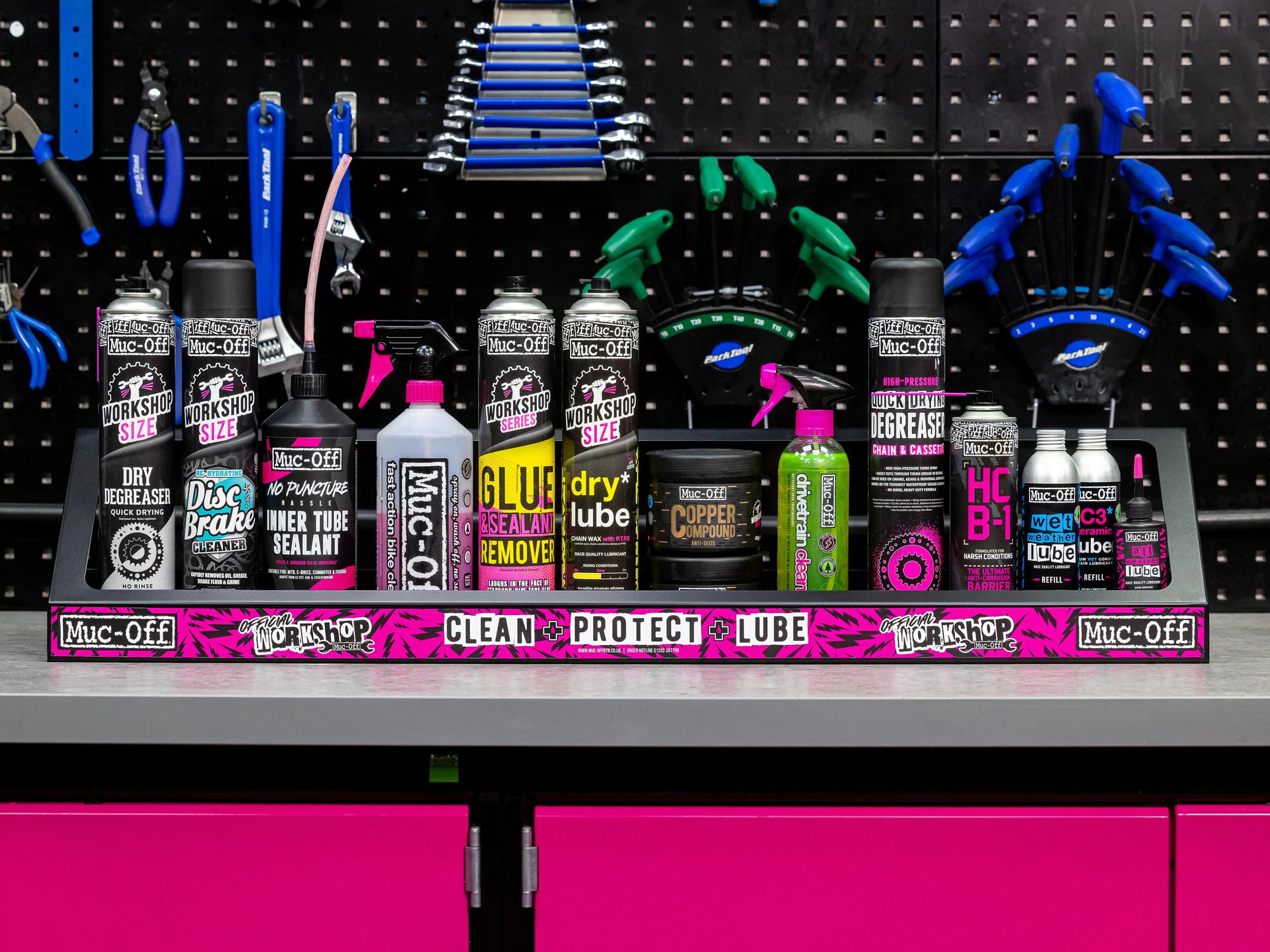 Row of various Muc-Off bike maintenance sprays and lubricants on a workbench with tools hanging on a pegboard background