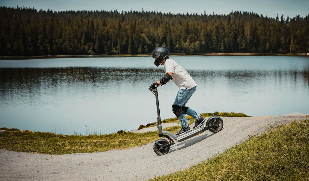 A person wearing a helmet and protective gear rides an electric scooter up a small hill on a path beside a lake, with a forest of tall trees in the background under a clear sky.