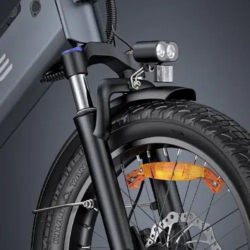 Close-up of a bicycles front wheel, showing the suspension fork, disc brake, thick tire, orange reflector, and a mounted dual headlight above the fender, with part of the frame in the background.