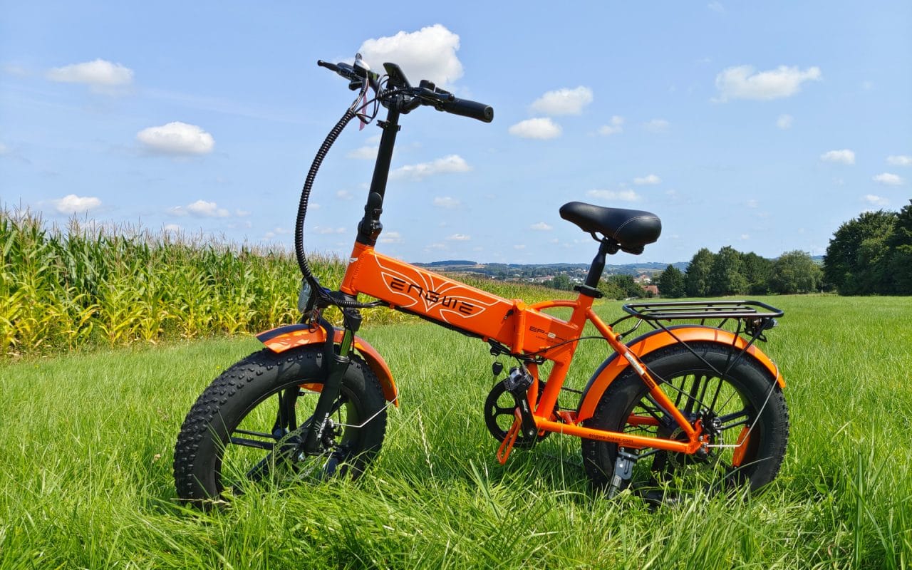 Orange electric bike with fat tires parked on green grass field under blue sky with scattered clouds