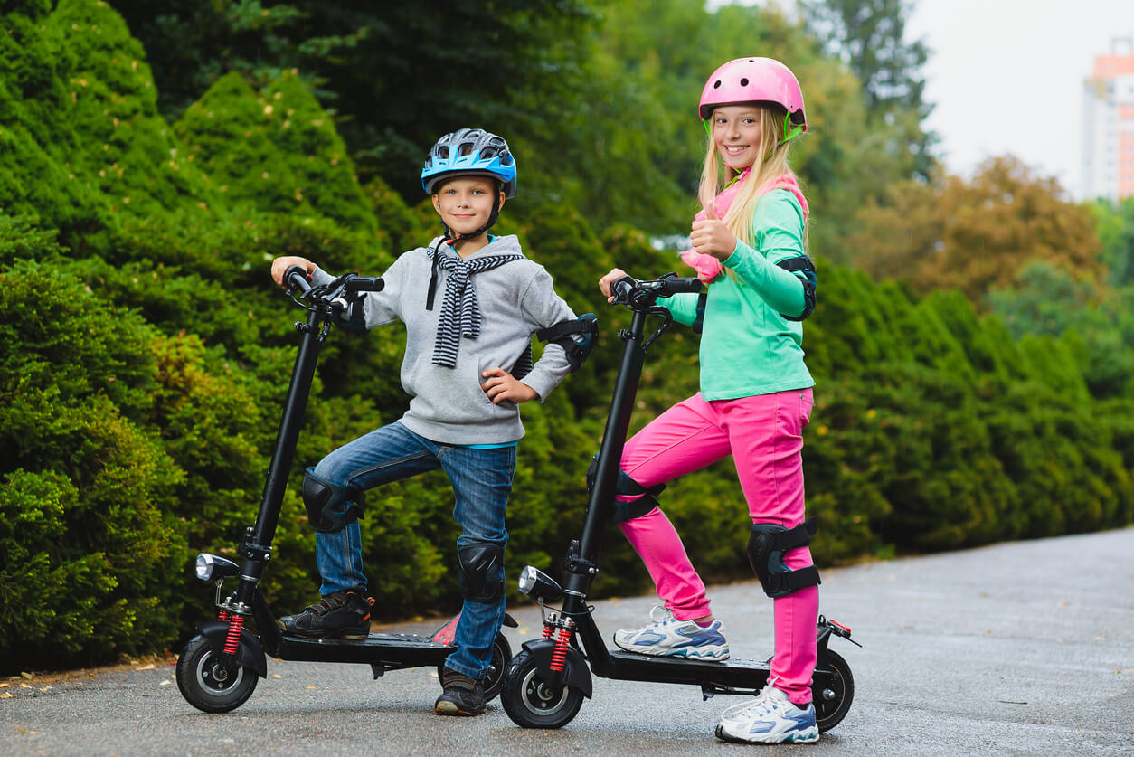 Two children wearing helmets standing with electric scooters on a path surrounded by green bushes outdoors