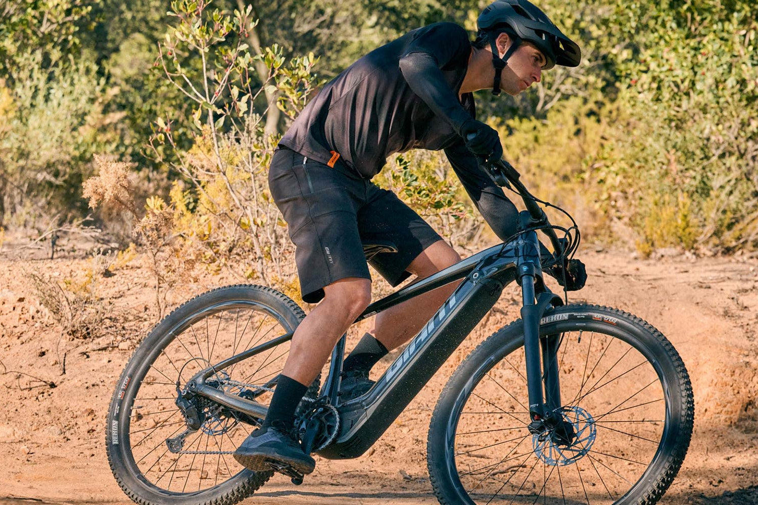 Man wearing black helmet and black cycling clothes riding a black mountain bike on a dirt trail outdoors