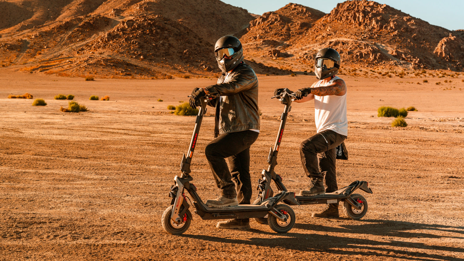 Two people wearing helmets and protective gear ride electric scooters across a dry, rocky desert landscape with mountains in the background.