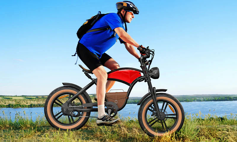 A man wearing a helmet, sunglasses, blue shirt, black shorts, and a backpack rides a red and black electric bicycle along a grassy path near a body of water on a clear, sunny day.