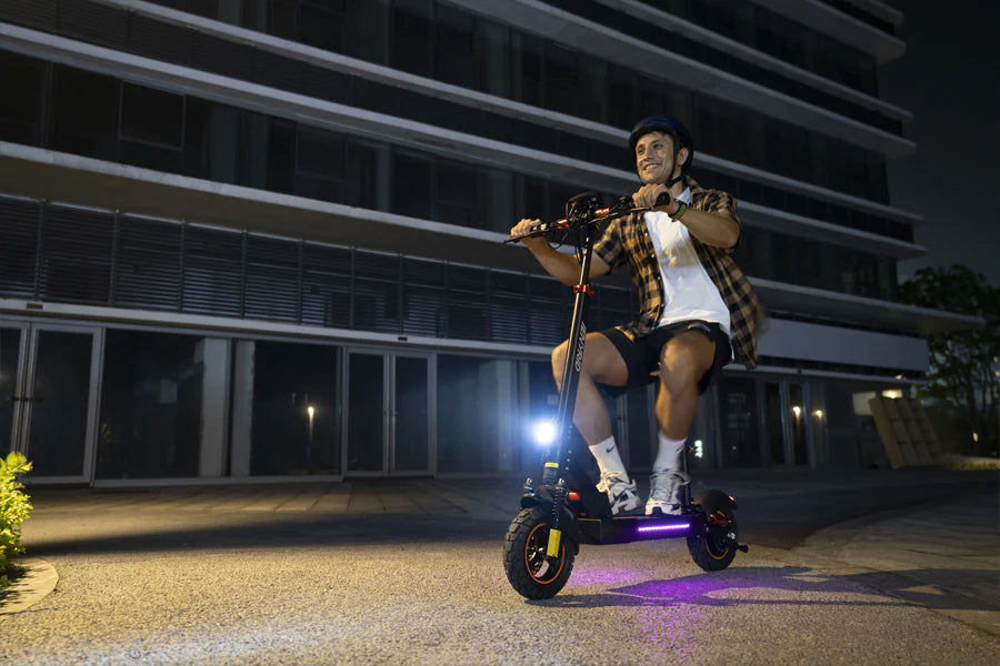 A person wearing a helmet rides an electric scooter at night in front of a modern building, illuminated by the scooters front light and ground lights.