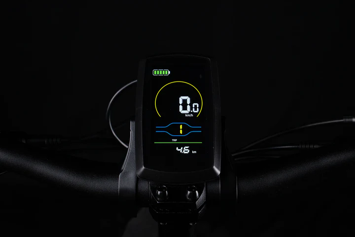 A close-up of an electric bike digital display shows speed at 0 km/h, a battery icon with four bars, a yellow circular outline, and other indicators against a black background. Handlebars are partially visible.