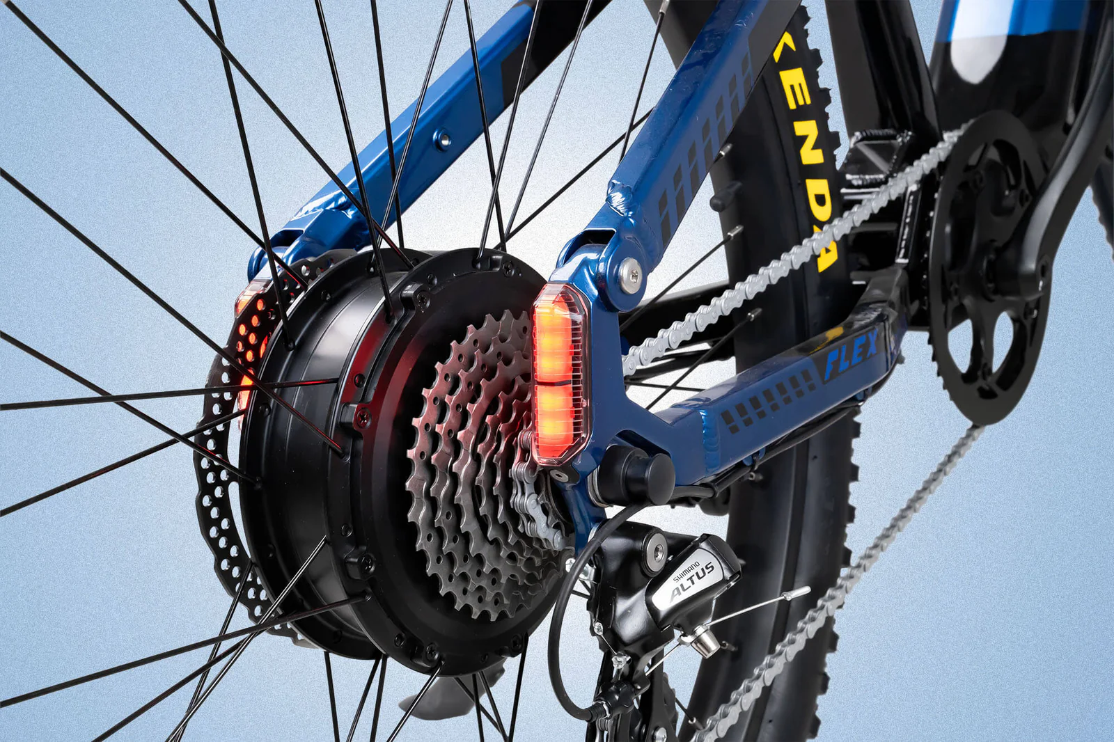 Close-up of a blue bicycle’s rear wheel showing the gear cassette, derailleur, disc brake, and a glowing red rear light integrated into the frame near the wheel spokes.