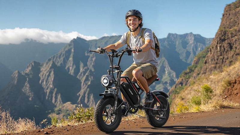 A smiling man wearing a helmet and backpack rides an electric bike on a mountain road, with a scenic backdrop of rugged mountains and a clear blue sky.