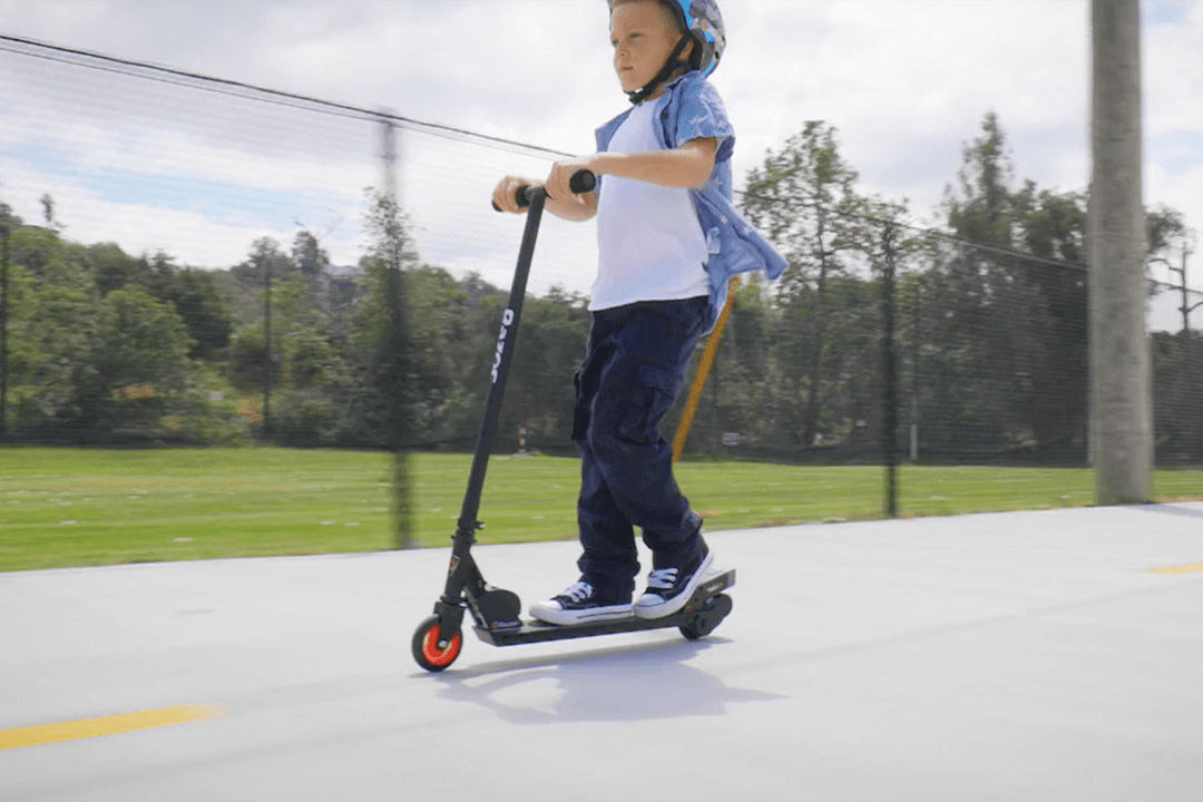 Young boy wearing a helmet riding a black electric scooter on a paved path with trees in the background
