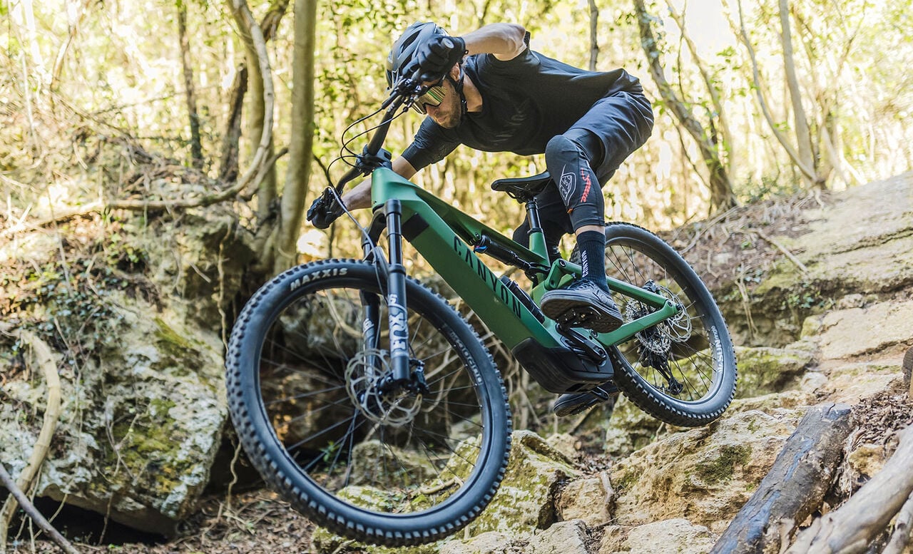 A person wearing a helmet and cycling gear rides a green mountain bike down a rocky, steep trail in a wooded area, navigating uneven terrain with focus and skill.