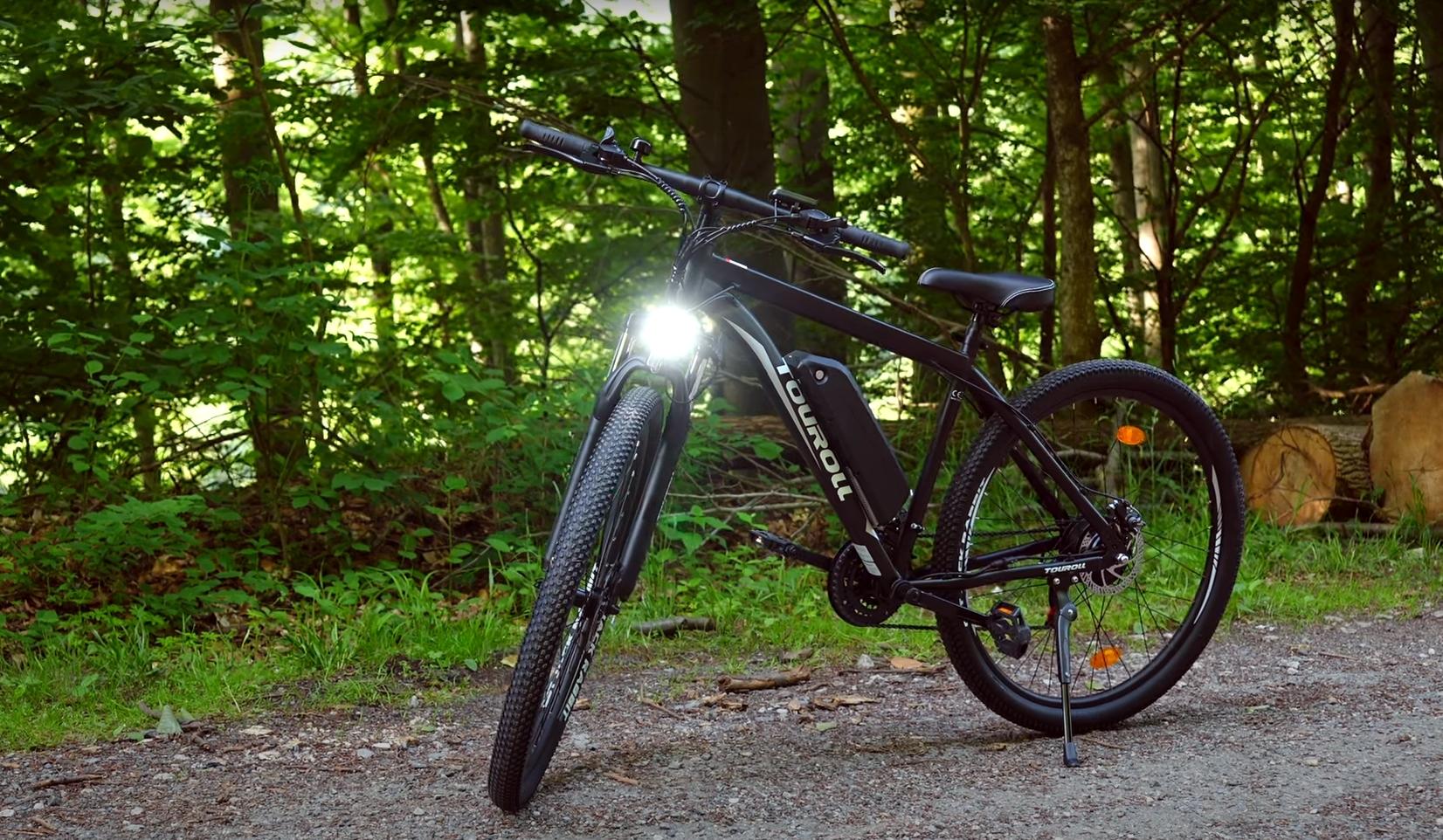 Black Currie electric bike with front light on gravel path surrounded by green forest and logs in background